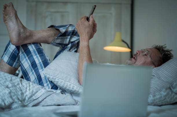 Homme de cinquante ans, en pyjama dans son lit, avec un téléphone portable et un ordinateur